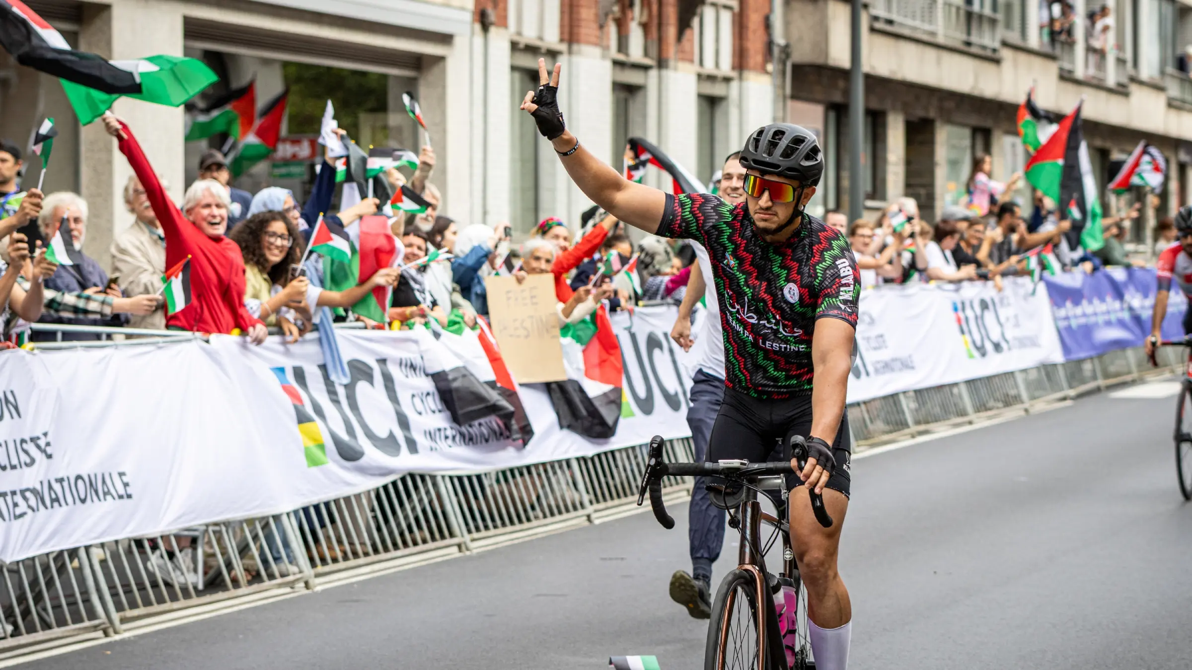 George Qadado cycling with Palestinian supporters waving flags at a UCI event